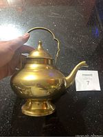 Top front angle showing the rounded brass teapot with its hinged lid and arched handle held by a hand.
