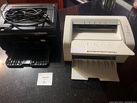 Photo showing two desktop laser printers, a black HP LaserJet P1102w and a white Samsung ML-2165 with cables on the HP printer.