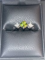 Close-up photo showing a silver ring with a central green square stone and two smaller diamond-colored square stones set on either side.