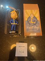 Vault Boy bobblehead in plastic display and Vault Boy Bobblehead labeled box side by side on reflective surface, with auction lot number card.