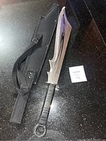 Custom fantasy sword with broad, uniquely shaped blade and black tribal pattern, placed on a black granite surface next to its black nylon carrying case with shoulder strap.