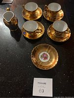 Overview of 4 gold-tone teacups, 6 saucers, and creamer on dark surface.