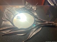 Full view of round mirror with decorative leather-like leaf and stem frame on black surface