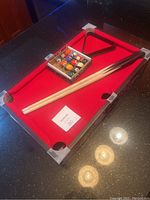 Miniature pool table showing red felt surface with two wooden cue sticks and black triangle rack placed on top.