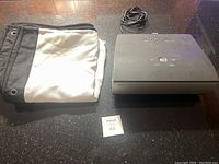 Photo showing the folded projector screen, the Sanyo SD projector with buttons on top, and power cable on a dark surface.