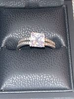 Front view of silver-colored ring with large square-cut diamond-colored stone on textured black cushion within a box.
