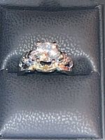 Close-up of silver toned skull ring with prominent diamond-colored center stone and smaller stones on the band, placed in a black ring box.