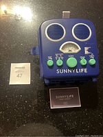 Front view of blue Sunnylife beach radio showing analog dial, speakers, and green control knobs.
