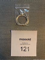 Silver toned thin necklace coiled in clear plastic bag on textured surface with lot number beneath.