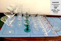 Photo showing 28 assorted bar glasses including martini glasses, sherbet glasses, champagne flutes, cocktail glasses, and tumblers laid out on a tablecloth.