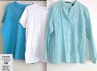 Three women's blouses displayed hanging side by side: blue and white short-sleeve jersey tops and a light aqua long-sleeve button-up blouse with floral embroidery.