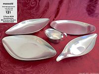 Five mid century modern Scandinavian stainless steel serving pieces arranged on burgundy fabric. Four serving trays with elongated leaf and oval shapes, one small creamer pitcher centrally placed. Serving trays are smooth with polished surfaces.