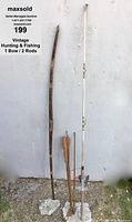 Full view of vintage wooden hunting bow and two fishing rods leaning against a wall with visible rocks on the ground.