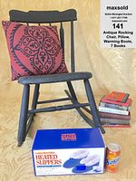 Antique black wooden rocking chair with decorative pillow on seat, heated slippers box, stack of 7 books and a nearby soda can for size comparison
