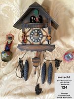 Front view of German cuckoo clock, brass goat bell, and red music box on fabric background with soda can for scale