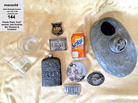 Top-down photo showing all items in lot including pewter flask, foot warmer, four belt buckles, two Baileys glasses, six coasters, and a soda can for size reference.
