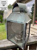 Front angled view of handcrafted copper lantern showing glass panels, hinged door, and metal framing with visible patina.