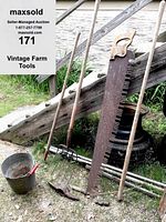 Full view of seven vintage farm tools propped against wooden fence, showing rusty 2-person saw, pitch forks, hoe/fork, hammer head, pickaxe, and long handle.