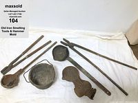 Overview of multiple old iron smelting tools including long handled scoops, rods, a bucket, and hammer mold displayed on a white cloth background.