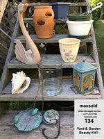 Entire lot displayed on wooden steps outdoors includes one ceramic swan planter, floral ceramic planter, blue metal lantern bird feeder, clear glass hanging planter, terracotta pot with openings, white plastic hanging planter, and a large sea shell. Soda can for size reference only.