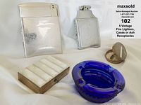 5 vintage smoking items: 2 silver cigarette case and lighter combos, 1 mother-of-pearl case, 1 small purse ashtray, and 1 blue glass ashtray, arranged on white cloth.