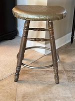 Full front-side view of antique wooden stool showing turned legs, metal ring footrest, and padded seat with recovered fabric cover.