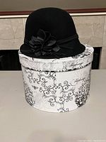 Black wool cloche hat displayed on a decorative round hat box with black script and floral patterns.
