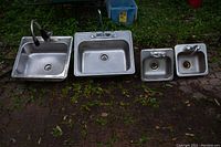 Four stainless steel sinks arranged on outdoor surface, showing two larger and two smaller sinks each with faucets attached.