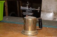 Photo showing front angled view of the brass torch highlighting the fuel tank, handle and metal burner with valves.