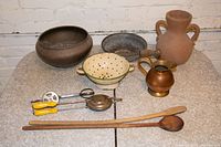 Overview of antique kitchenware including ceramic jug with pennies, metal bowls, enamel strainer, hand crank egg beater, wooden utensils, and copper/brass pitcher