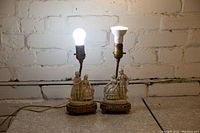 Pair of antique lamp bases with ceramic figurines and lit bulbs on a table against a white brick wall.