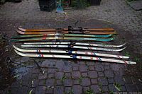 Four pairs of vintage skis of different styles and colors arranged on cobblestone ground, showing overall condition and variety.
