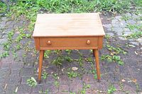 Front view of a wooden Mid-Century Modern bedside table with one drawer and tapered legs on an outdoor paved ground.