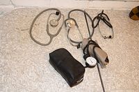 Photo showing all items: three stethoscopes placed on floor next to a blood pressure pump with attached cuff and gauge, plus black case.