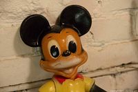 Close-up of Mickey Mouse rubber toy head and upper body against white brick wall.