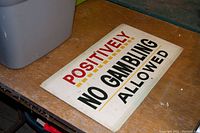 Photo of the vintage rectangular sign on a wooden surface showing full text "POSITIVELY NO GAMBLING ALLOWED" in red, black, and yellow colors.