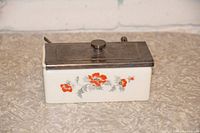 Closed view of the vintage white ceramic serving container with orange floral decoration and a rectangular metal sliding lid with knob.