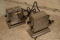 Photo showing two vintage chrome electric toasters with fabric covered cords on a surface against a brick backdrop.