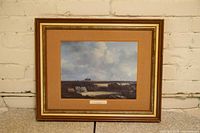 Framed print hanging on a white brick wall showing a landscape scene with sky, fields and small buildings, matted in tan with a wooden and gold frame.