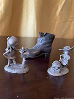Three pewter figurines displayed on a table including a girl by mailbox, a scarecrow, and a shoe figurine
