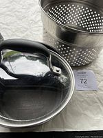 Photo showing the Analon pot with glass lid and the stainless steel strainer basket placed near each other on a light fabric surface.