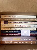 Top-down photo showing spines of 10 cookbooks in a cardboard box, titles and years visible.