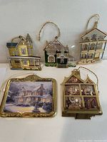 Front view of five ornaments arranged on white surface showing detailed house shapes and Thomas Kinkade plaque style ornament