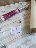 Photo showing both the Mirage wallcoverings roll and the Con-Tact paper lying on a floral patterned surface with a lot tag.