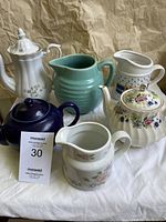 Full set of six ceramic and porcelain items showing variation in color, styles, and shapes including a white tall coffee pot, green pitcher, duckling pitcher, Sadler and cobalt blue teapots, and a floral pitcher.
