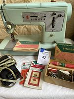 Front left angled view of the Singer Model 338 sewing machine with original instruction manual, box of Singer needles, pedal, and machine-related accessories.