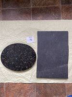 Both rock slabs shown on a textured beige surface, the oval is polished black with white flecks, and the rectangular is solid matte dark gray.