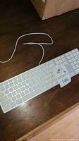 Full view of Apple wired keyboard with attached USB cable lying on wooden surface, showing numeric keypad and white keys.