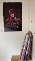Poster featuring Daredevil in red costume with target like graphic behind head, mounted on wall with tubes on a table below