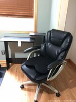 Photo showing black leather office chair on rollers next to the black computer desk under window with wood blinds.